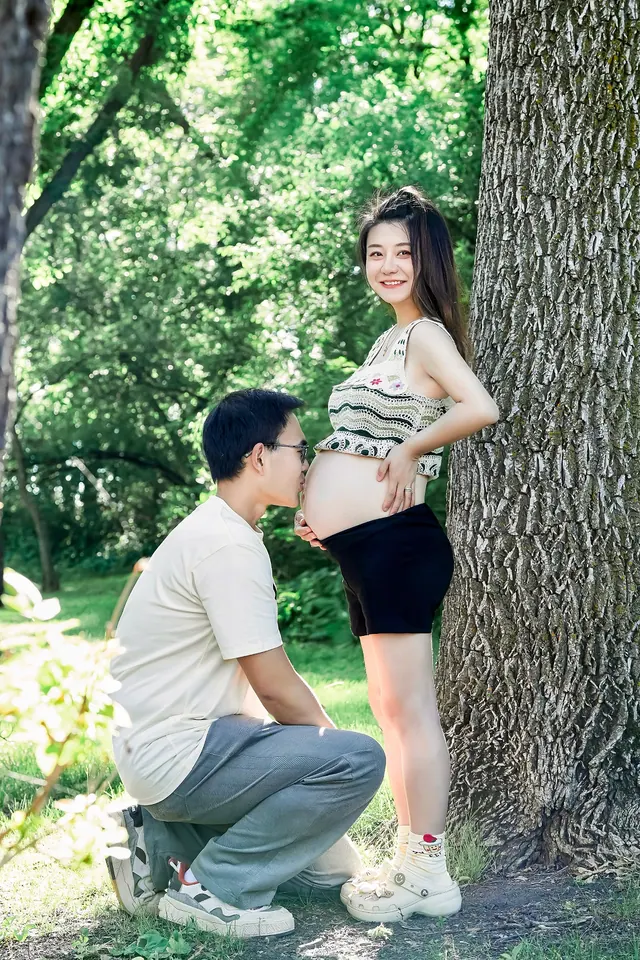 大树旁，准妈妈站着微笑，另一位大人蹲在身边轻触孕肚